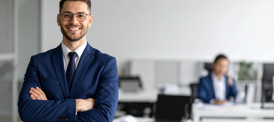 man with glasses in blue business suit in an office blurred out behind him with a coworker sitting at a desk